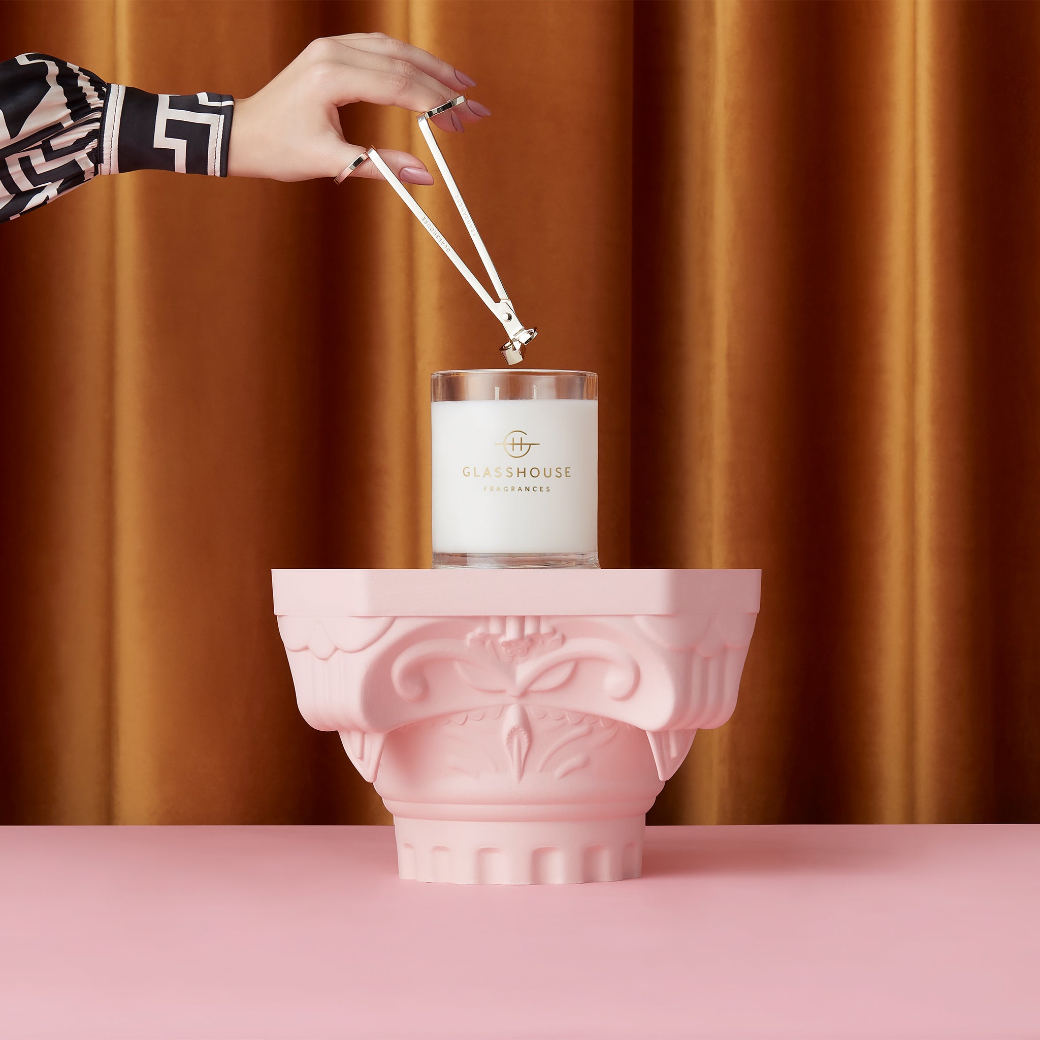 Hand holding Glasshouse Fragrances Wick trimmer over Soy Candle sitting on a pink stand with rust colour curtain background